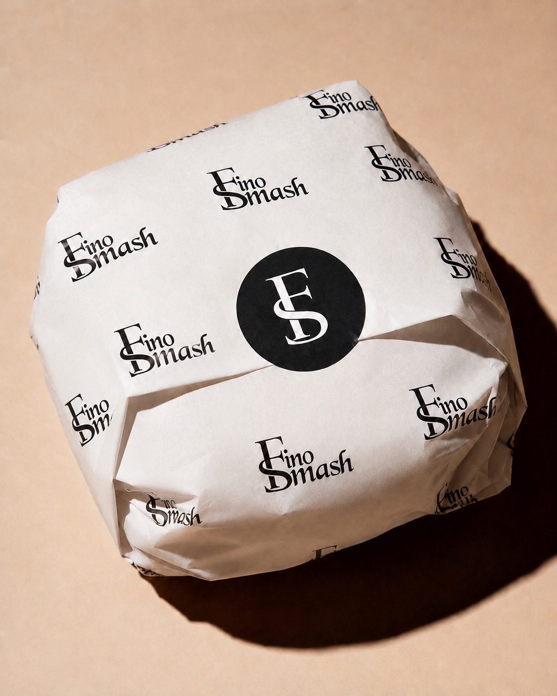 FinoSmash campaign: a burger wrapped in FinoSmash printed paper, sealed with the black FS seal, shot from above on sand